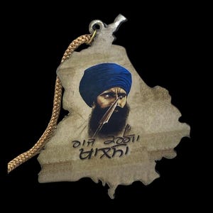 May include: A pendant in the shape of a map, featuring a portrait of a man with a blue turban and beard. The pendant has a gold-colored cord attached for wearing. The image has text in a foreign language.