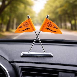 Può includere: Due piccole bandiere arancioni con simboli neri sono incrociate e montate su un supporto argentato. Le bandiere sono esposte sul cruscotto di un'auto, con uno sfondo sfocato di una strada fiancheggiata da alberi.