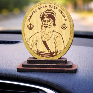 May include: Wooden car dashboard decoration with an illustration of Shaheed Baba Deep Singh Ji. The round emblem has gold glitter edging and is mounted on a tiered wooden base. The text "SHAHEED BABA DEEP SINGH JI" is inscribed around the image.