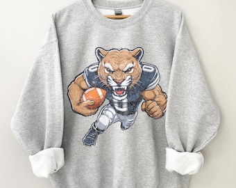 Könnte beinhalten: Ein hellgraues Sweatshirt mit einer Illustration eines Cougars in Football-Uniform, der einen orangefarbenen Football hält. Der Cougar hat einen aggressiven Gesichtsausdruck. Das Sweatshirt hat einen Rundhalsausschnitt und lange Ärmel.