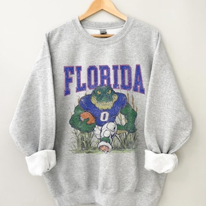 May include: Grey sweatshirt featuring a vintage-style graphic of a green alligator holding a football. The text "FLORIDA" is printed in purple above the alligator.