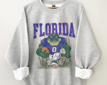 May include: Gray sweatshirt with a vintage-style graphic of a green alligator holding a football. The text "FLORIDA" is printed in purple above the alligator.