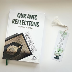 May include: A white book titled "Qur'anic Reflections" with the subtitle "Daily Verse for 30 Days." The book features a decorative cover and a green ribbon bookmark. A matching bookmark with Arabic text and a tassel is also present.