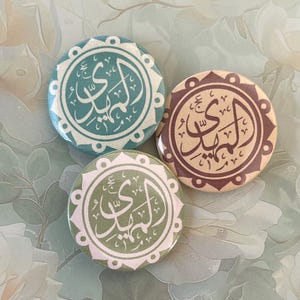 May include: Three round buttons with Arabic calligraphy in teal, olive green, and brown. Each button features a white decorative border and text. The buttons are arranged on a floral background.
