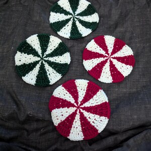 May include: Four crocheted peppermint candy coasters in red, green, and white. The coasters are round and have a swirl design.