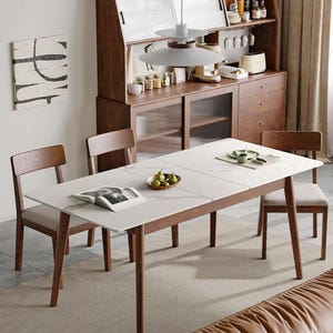 May include: A rectangular dining table with a white marble-patterned top and four wooden legs, accompanied by four wooden chairs with light-colored upholstered seats. An open book and a small bowl of green pears sit on the table surface.