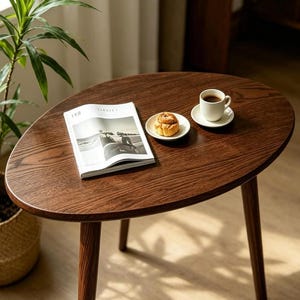 Puede incluir: Una mesa de centro de madera de color marr&oacute;n oscuro con una superficie ovalada y tres patas c&oacute;nicas. Sobre la mesa hay una revista abierta, un peque&ntilde;o plato de cer&aacute;mica blanca con un pastel y una taza de caf&eacute; blanca sobre su platillo.