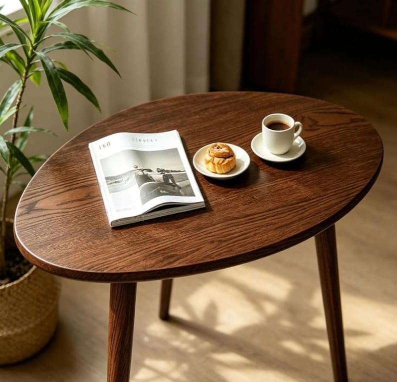 Puede incluir: Una mesa de centro de madera de color marr&oacute;n oscuro con una superficie ovalada y tres patas c&oacute;nicas. Sobre la mesa hay una revista abierta, un peque&ntilde;o plato de cer&aacute;mica blanca con un pastel y una taza de caf&eacute; blanca sobre su platillo.