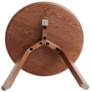 Puede incluir: Una mesa auxiliar redonda de madera vista desde abajo, que muestra el tablero circular con una veta de madera visible y tres patas anguladas unidas al centro con tornillos negros y almohadillas de fieltro gris claro en los pies.