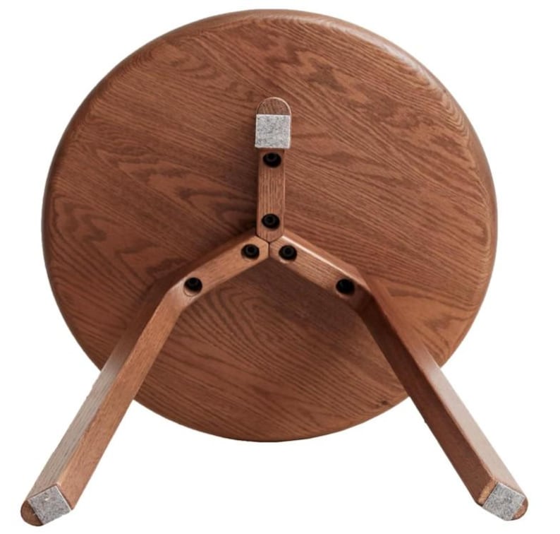 Puede incluir: Una mesa auxiliar redonda de madera vista desde abajo, que muestra el tablero circular con una veta de madera visible y tres patas anguladas unidas al centro con tornillos negros y almohadillas de fieltro gris claro en los pies.