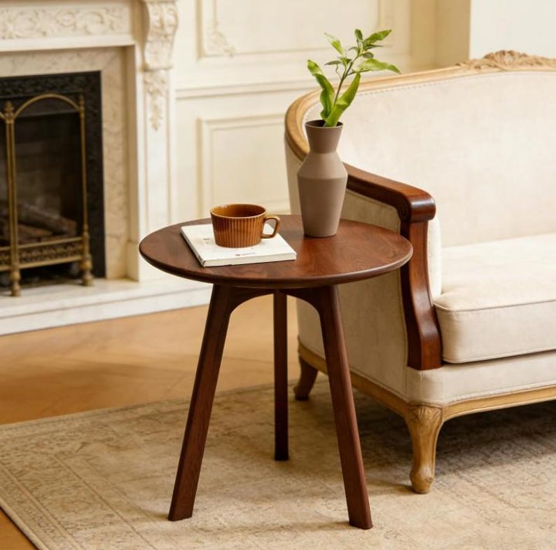 Puede incluir: Una mesa auxiliar redonda de madera marr&oacute;n oscuro con tres patas c&oacute;nicas. Sobre la mesa hay un libro rectangular blanco, una taza de cer&aacute;mica marr&oacute;n acanalada y un jarr&oacute;n alto de color beige mate con una rama de planta verde.