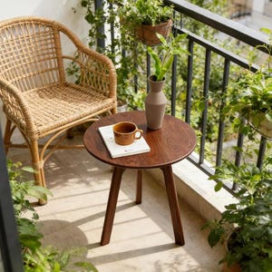 Puede incluir: Una mesa auxiliar redonda de madera con tres patas c&oacute;nicas, que presenta una taza de cer&aacute;mica marr&oacute;n claro y un libro blanco sobre su superficie. Junto a la mesa hay un sill&oacute;n de rat&aacute;n trenzado con respaldo alto y reposabrazos curvos en un balc&oacute;n.