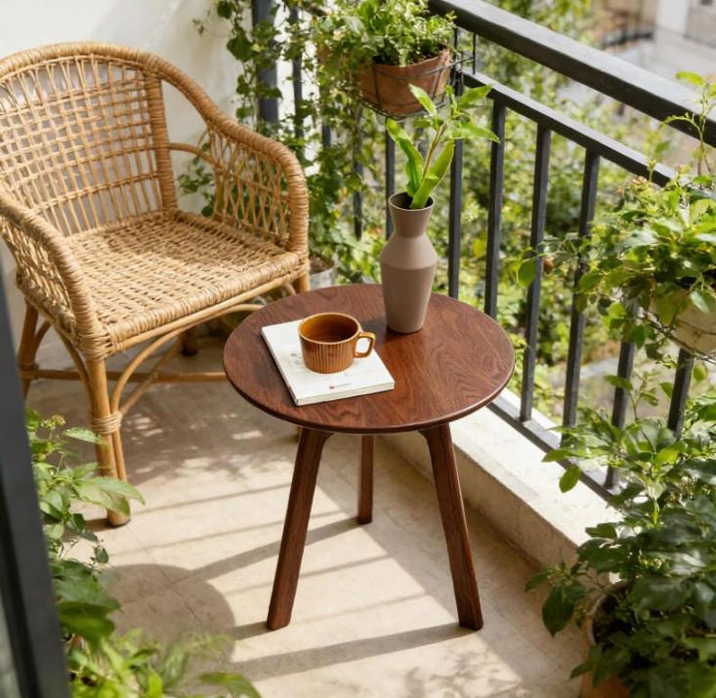 Puede incluir: Una mesa auxiliar redonda de madera con tres patas c&oacute;nicas, que presenta una taza de cer&aacute;mica marr&oacute;n claro y un libro blanco sobre su superficie. Junto a la mesa hay un sill&oacute;n de rat&aacute;n trenzado con respaldo alto y reposabrazos curvos en un balc&oacute;n.