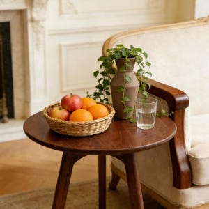 Puede incluir: Una mesa auxiliar redonda de madera sostiene una cesta tejida llena de dos manzanas rojas y dos naranjas. Junto a la cesta hay un jarr&oacute;n marr&oacute;n mate con una planta verde colgante y un vaso de cristal transparente lleno de agua.