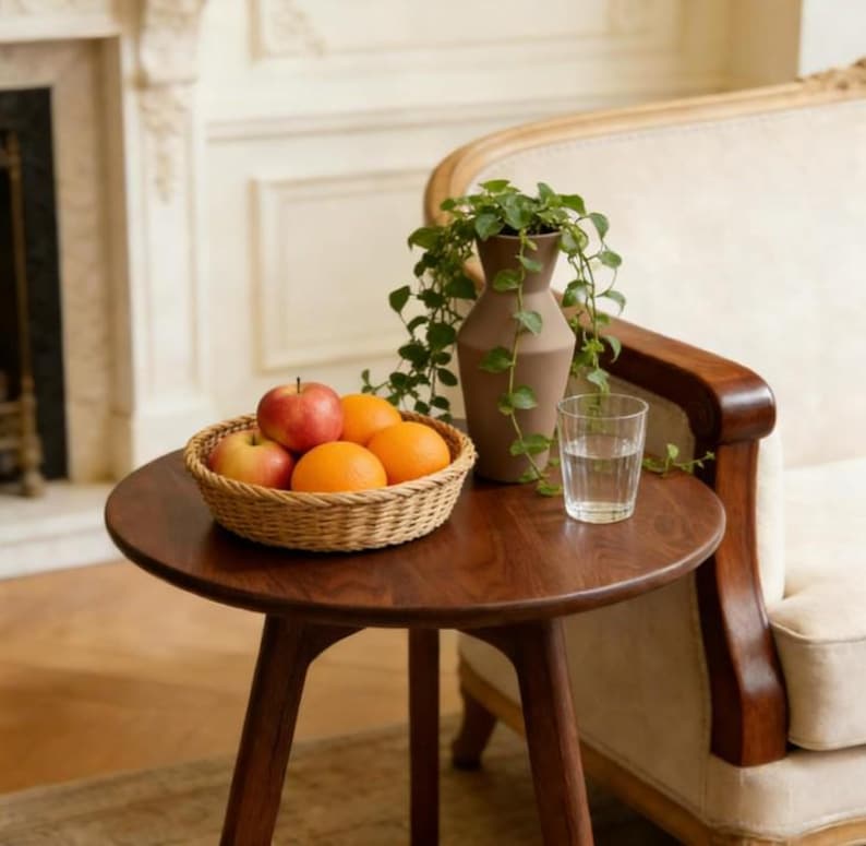 Puede incluir: Una mesa auxiliar redonda de madera sostiene una cesta tejida llena de dos manzanas rojas y dos naranjas. Junto a la cesta hay un jarr&oacute;n marr&oacute;n mate con una planta verde colgante y un vaso de cristal transparente lleno de agua.