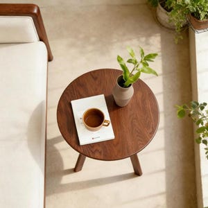 Puede incluir: Una mesa auxiliar redonda de madera oscura con tres patas, que presenta un libro blanco con una taza de cer&aacute;mica marr&oacute;n y un peque&ntilde;o jarr&oacute;n claro con una planta verde, situada junto a un sof&aacute; de color crema con estructura de madera.