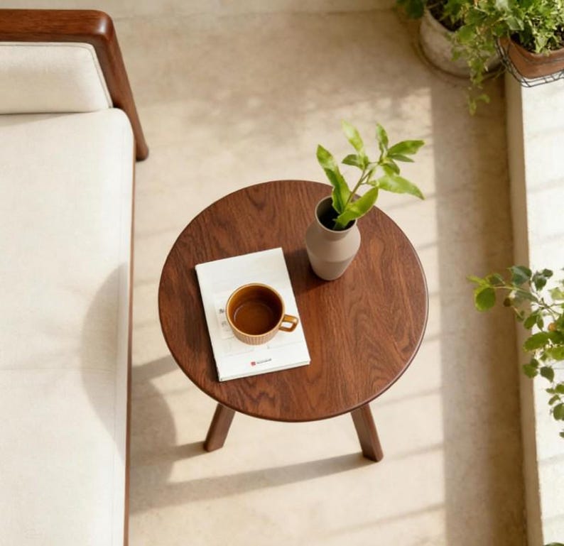 Puede incluir: Una mesa auxiliar redonda de madera oscura con tres patas, que presenta un libro blanco con una taza de cer&aacute;mica marr&oacute;n y un peque&ntilde;o jarr&oacute;n claro con una planta verde, situada junto a un sof&aacute; de color crema con estructura de madera.