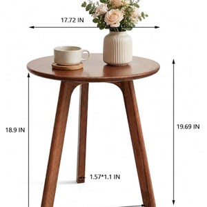 Puede incluir: Una mesa auxiliar redonda de madera con tres patas inclinadas, que mide 45 cm de di&aacute;metro y 50 cm de altura. La mesa tiene un acabado marr&oacute;n claro y sostiene un jarr&oacute;n de cer&aacute;mica beige con flores y una taza beige sobre un posavasos.