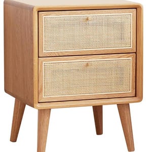 May include: A light brown wooden nightstand featuring two drawers with woven cane panels. The piece has rounded corners, a smooth finish, and four tapered legs. Each drawer is equipped with a small, circular, light-colored metal knob.