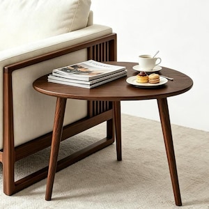 Puede incluir: Una mesa auxiliar de madera marr&oacute;n oscuro con una superficie triangular redondeada y tres patas c&oacute;nicas. La mesa sostiene una pila de revistas, una taza de caf&eacute; de cer&aacute;mica blanca sobre un platillo y un plato peque&ntilde;o con dos pasteles redondos.