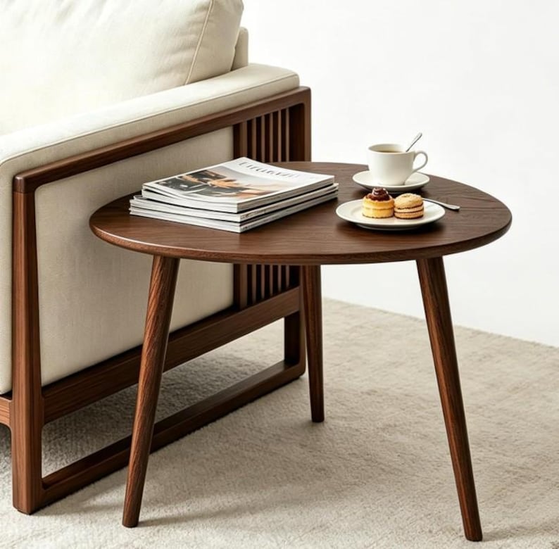 Puede incluir: Una mesa auxiliar de madera marr&oacute;n oscuro con una superficie triangular redondeada y tres patas c&oacute;nicas. La mesa sostiene una pila de revistas, una taza de caf&eacute; de cer&aacute;mica blanca sobre un platillo y un plato peque&ntilde;o con dos pasteles redondos.