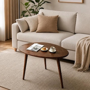 Puede incluir: Una mesa de centro de madera de color marr&oacute;n oscuro con una superficie ovalada y tres patas c&oacute;nicas. Sobre la mesa hay un libro abierto, un plato peque&ntilde;o blanco con un pastel y una taza de cer&aacute;mica blanca sobre un platillo a juego.