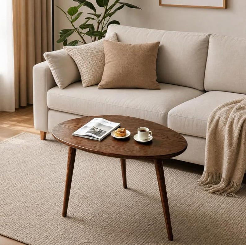 Puede incluir: Una mesa de centro de madera de color marr&oacute;n oscuro con una superficie ovalada y tres patas c&oacute;nicas. Sobre la mesa hay un libro abierto, un plato peque&ntilde;o blanco con un pastel y una taza de cer&aacute;mica blanca sobre un platillo a juego.