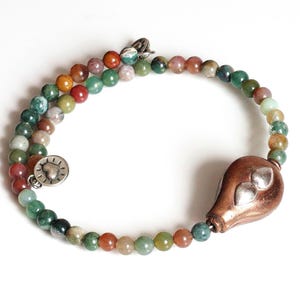 May include: A double-strand bracelet featuring colorful, round beads in shades of green, red, orange, and brown. The bracelet has a copper-colored teardrop-shaped charm with silver accents and a silver heart-shaped clasp.