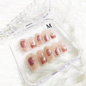 May include: A clear plastic case with a set of 10 false nails. The nails are a light pink color with a gold design and a pink accent. The case has a label that says "M".
