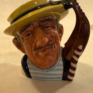 Puede incluir: Taza de cerámica con forma de cabeza de hombre, con sombrero amarillo y camisa a rayas. El asa es un remo de góndola. El hombre tiene bigote y expresión seria. Artículo decorativo.