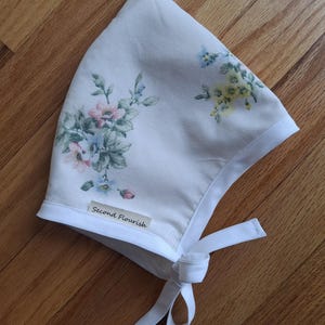 May include: A white bonnet with a floral pattern in pink, blue, and yellow. The bonnet has a white trim and ties, and a small label that reads "Second Flourish".