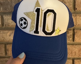 Può includere: Cappellino da camionista blu e bianco con il numero "10" in nero e glitter dorati. Una grafica di pallone da calcio è sul lato sinistro, con stelle glitterate dorate sul cappello.