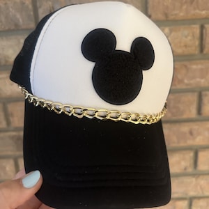 May include: Black and white baseball cap featuring a gold chain and a black Mickey Mouse silhouette on the front. The cap has a mesh back and a curved brim. The hat is a stylish accessory.