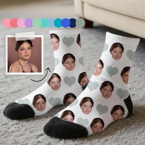 Peut inclure: Une paire de chaussettes blanches avec un motif répété de visages de femme et de coeurs gris. Les chaussettes ont des talons et des orteils noirs. Une photo de la femme est collée en haut à gauche.