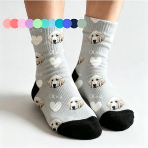 May include: Light grey socks with a repeating pattern of a golden retriever's face, white hearts, and the name "Olivia". The socks have black heels and toes.