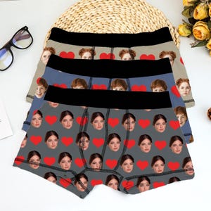 Puede incluir: Tres pares de bóxers con patrones personalizados de caras y corazones. Los bóxers tienen una cintura negra y son de color gris, azul y beige. El par gris tiene un patrón de caras y corazones rojos. Los pares azul y beige también tienen caras y corazones.