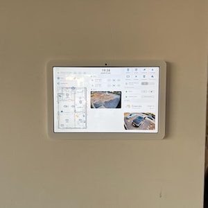 Può includere: Un tablet bianco montato su una parete mostra un'interfaccia di sistema di sicurezza domestica. Lo schermo mostra una piantina, un feed video in diretta di un vialetto e una lettura della temperatura di 23,3 °C. L'ora è le 19:34.