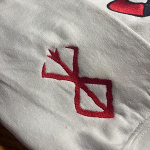 May include: White long-sleeve shirt with a red embroidered design on the sleeve. The design is a stylized symbol. A red and black Nike logo is visible in the upper right corner.