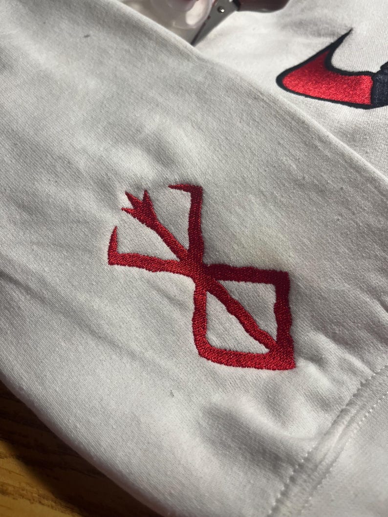 May include: White long-sleeve shirt with a red embroidered design on the sleeve. The design is a stylized symbol. A red and black Nike logo is visible in the upper right corner.
