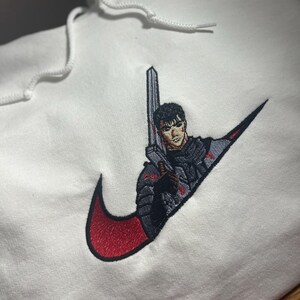 May include: White hoodie with a red and black embroidered design. The design features a character holding a sword, integrated into a swoosh logo. The hoodie has a drawstring hood.