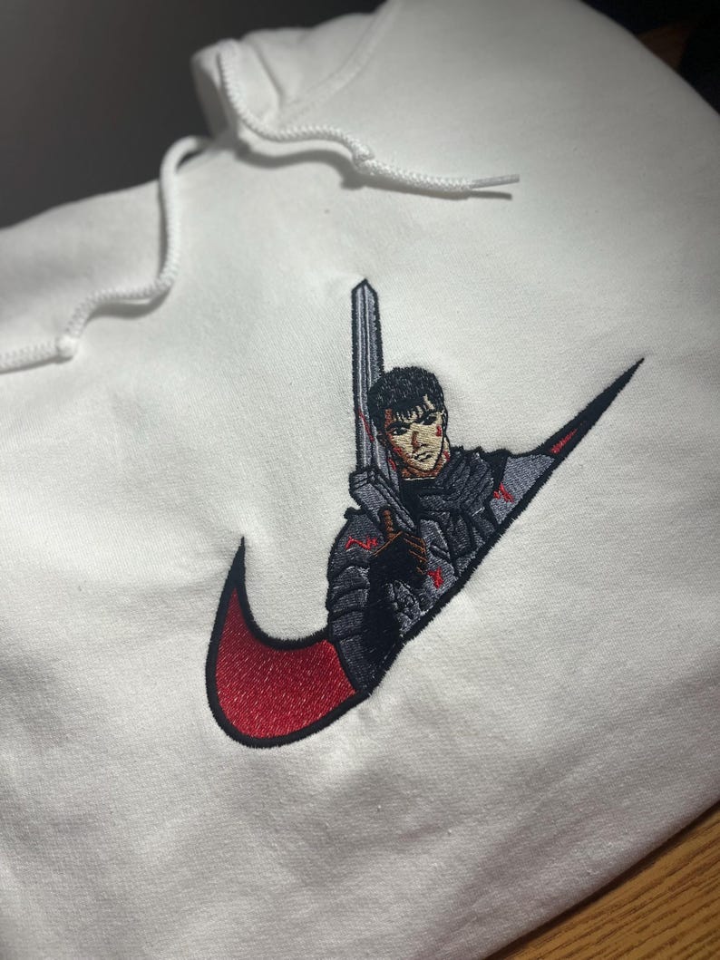 May include: White hoodie with a red and black embroidered design. The design features a character holding a sword, integrated into a swoosh logo. The hoodie has a drawstring hood.