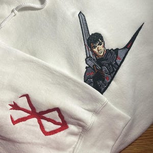 May include: White hoodie with embroidered artwork. The design features a character holding a sword, with red accents. The sleeve displays a red emblem. The hoodie is made of a soft material.