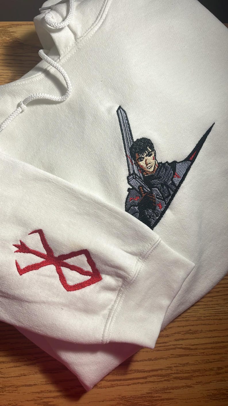 May include: White hoodie with embroidered artwork. The design features a character holding a sword, with red accents. The sleeve displays a red emblem. The hoodie is made of a soft material.