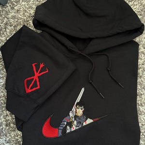 May include: Black hooded sweatshirt with a red and white embroidered design. The design features a character holding a sword, integrated into a red swoosh. The left sleeve has a red symbol.