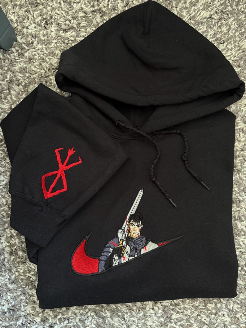 May include: Black hooded sweatshirt with a red and white embroidered design. The design features a character holding a sword, integrated into a red swoosh. The left sleeve has a red symbol.