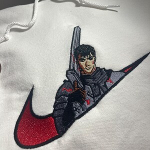 May include: White hoodie with a red and black embroidered design. The design features a character holding a large sword, integrated into a swoosh logo. The hoodie has a drawstring hood.