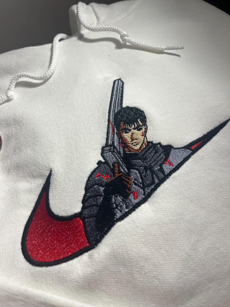May include: White hoodie with a red and black embroidered design. The design features a character holding a large sword, integrated into a swoosh logo. The hoodie has a drawstring hood.