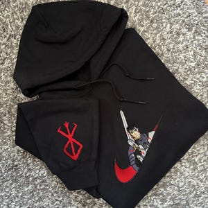 May include: Black hooded sweatshirt featuring a red and white embroidered anime character design within a red swoosh. The left sleeve has a red embroidered symbol. The sweatshirt has a drawstring hood.