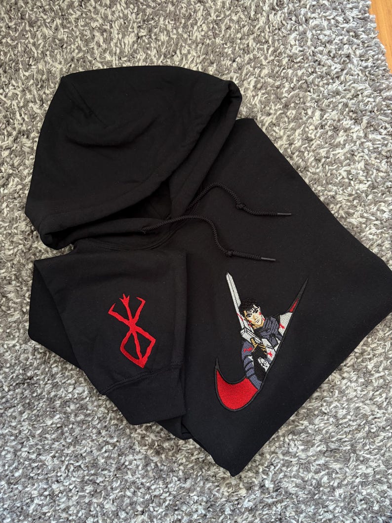 May include: Black hooded sweatshirt featuring a red and white embroidered anime character design within a red swoosh. The left sleeve has a red embroidered symbol. The sweatshirt has a drawstring hood.