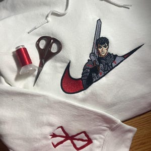 May include: White hoodie with a red and black embroidered design featuring a character with a sword. The design is incorporated into a swoosh logo. A red thread spool and scissors are also visible.